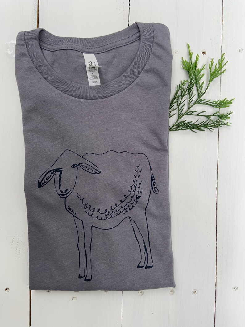 May include: A gray t-shirt with a black and white illustration of a sheep. The sheep is drawn in a simple, stylized way, with a curly tail and a friendly face.