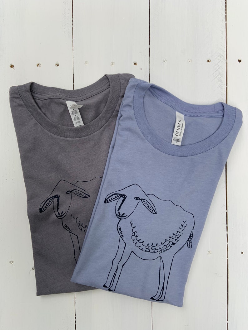 May include: Two t-shirts with a simple black line drawing of a sheep on them. The shirts are a light gray and a light blue.