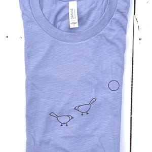 May include: A light blue t-shirt with a simple design of two birds and a circle. The shirt is made of a soft, comfortable fabric.