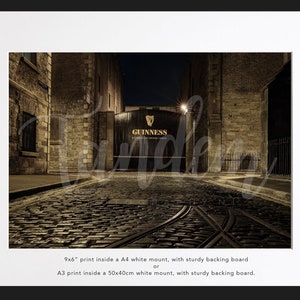 May include: A nighttime view of a cobblestone street leading to a large, dark wooden gate with the words "GUINNESS" printed in gold lettering. The gate is flanked by brick buildings.