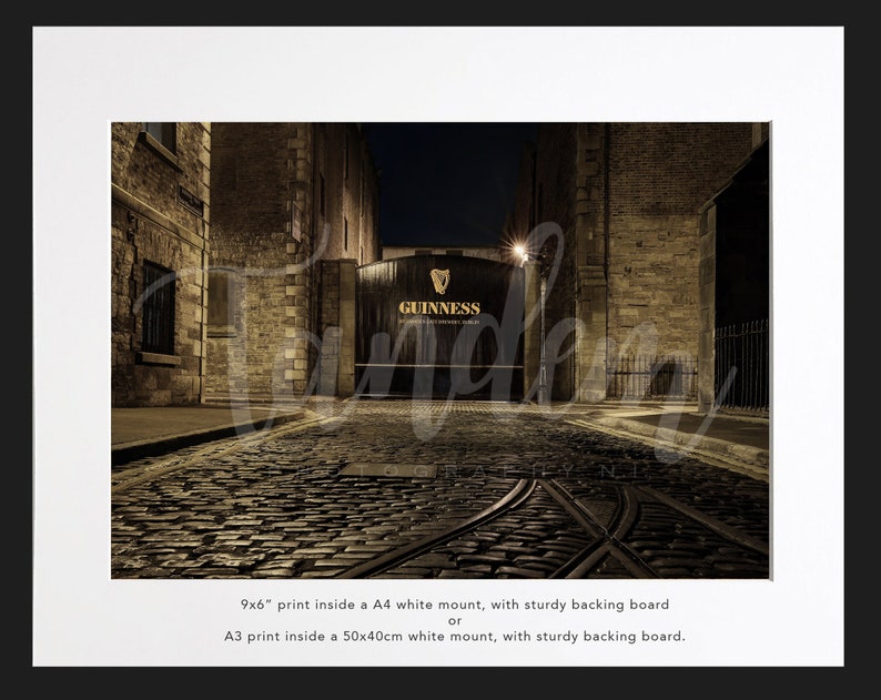 May include: A nighttime view of a cobblestone street leading to a large, dark wooden gate with the words "GUINNESS" printed in gold lettering. The gate is flanked by brick buildings.