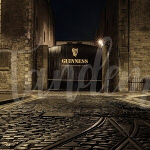 May include: A dark, narrow cobblestone street leads to a large wooden gate with the words "GUINNESS, ST. JAMES'S GATE BREWERS, DUBLIN" in gold lettering. The gate is lit by a street lamp.