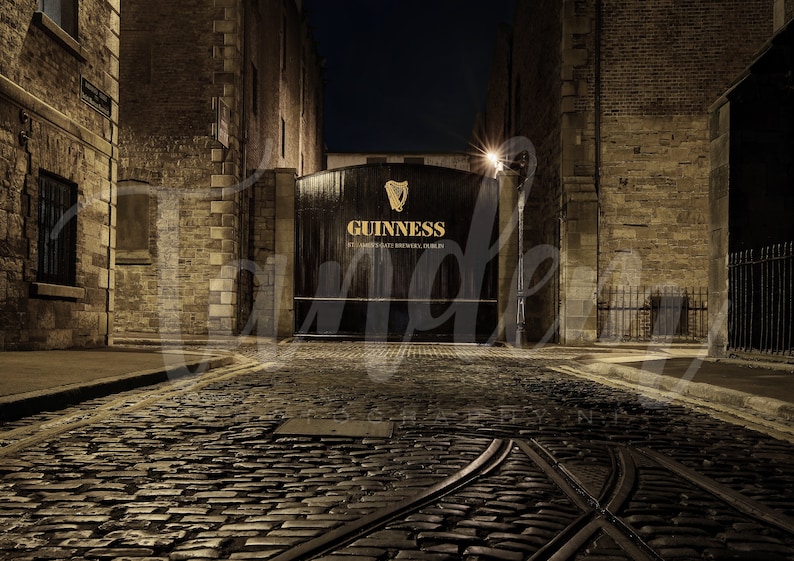 May include: A dark, narrow cobblestone street leads to a large wooden gate with the words "GUINNESS, ST. JAMES'S GATE BREWERS, DUBLIN" in gold lettering. The gate is lit by a street lamp.
