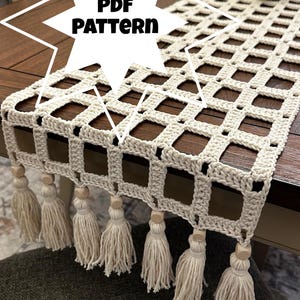 Op de afbeelding: Crèmekleurige gehaakte tafelloper met een rasterpatroon en kwastjes. De loper wordt gepresenteerd op een donkere houten tafel. De tekst "PDF PATTERN" staat in een sterburst-afbeelding.