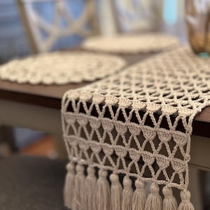 May include: A beige, open-weave crochet table runner with a geometric pattern and fringe tassels. The runner is displayed on a dark wood table, with matching placemats and chairs in the background. The tassels hang down from the edge of the table runner.