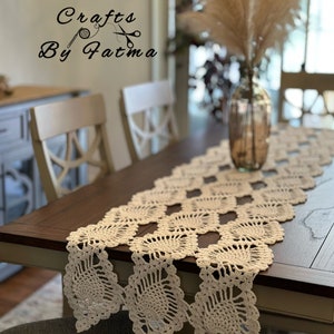 May include: Cream-colored, intricately crocheted table runner. The runner features a repeating geometric pattern with scalloped edges, displayed on a dark wooden table. The text "Crafts By Fatima" is visible in the upper left corner. A vase with dried pampas grass is in the background.