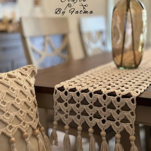 以下が含まれることがあります： クリーム色のかぎ針編みのテーブルランナー。スカラップエッジとタッセル付きで、濃い色の木製テーブルに飾られています。タッセルと木製ビーズが付いたお揃いの枕も見えます。画像には、装飾用のガラスの花瓶と「Crafts By Fatma」の文字も表示されています。