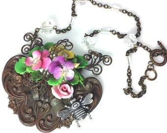 May include: A necklace featuring a large, ornate bronze-toned pendant with intricate scrollwork, adorned with a cluster of pink, purple, and yellow flowers with clear rhinestones, and a silver-toned bee charm, suspended from a bronze chain with clear beads.