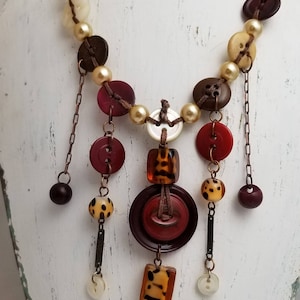 May include: A unique button necklace featuring a variety of button shapes and colors, including brown, red, and cream. The necklace also includes faux pearls and decorative beads, creating a layered, artistic design.