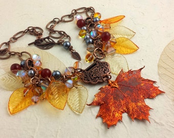 May include: A copper-coloured necklace with a fall theme. The necklace features a large maple leaf pendant, amber-coloured beads, and translucent yellow leaf charms. The chain is made of copper-coloured links. The necklace is set against a textured, off-white background.