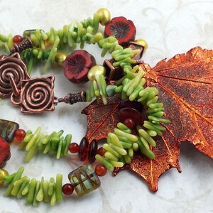 Op de afbeelding: Een handgemaakte ketting met een koperen sluiting en een groot koperen blad. De ketting bevat groene, rode en gouden kralen, samen met rode bloemvormige kralen. Het ontwerp roept een herfstthema op.