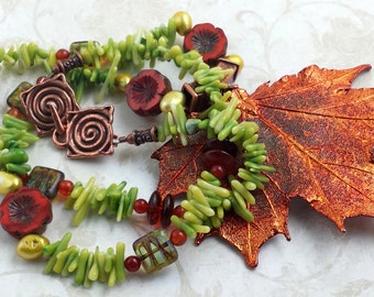 May include: A handcrafted necklace with a copper clasp and a large copper leaf. The necklace incorporates green, red, and gold beads, along with red flower-shaped beads. The design evokes an autumnal theme.