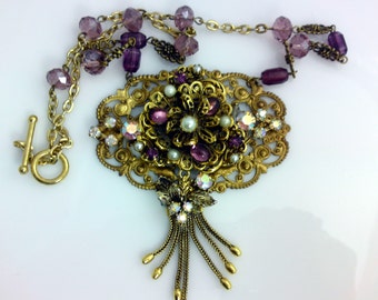 May include: A gold-tone necklace featuring an ornate filigree pendant adorned with purple cabochons, small white pearls, and iridescent rhinestones. The pendant includes a dangling tassel of gold chains and is attached to a chain of purple beads.