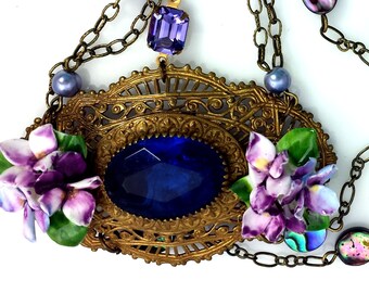 May include: A necklace featuring a central oval deep blue faceted stone set in an ornate brass filigree frame. Two clusters of purple and white ceramic flowers flank the stone. The piece includes purple beads, a rectangular purple stone, and chain links.