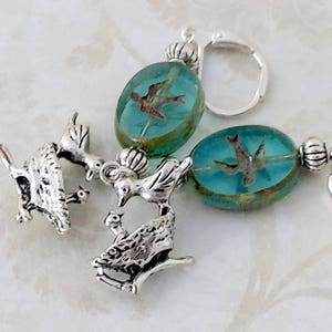 May include: Close-up of handmade earrings featuring silver bird charms and teal-colored glass beads with embedded bird designs. The earrings have a lever-back closure. The design is intricate and detailed, with a vintage aesthetic.