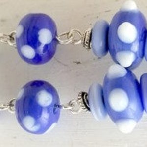 Puede incluir: Dos pares de pendientes hechos a mano con cuentas de vidrio. Cada pendiente presenta una combinación de cuentas azules y blancas con lunares, conectadas por cadenas de color plateado y detalles metálicos decorativos. Los pendientes están diseñados con una variedad de formas y tamaños de cuentas.