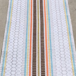 May include: A white woven rug with brown, orange, yellow, and blue stripes. The rug has a diamond pattern.