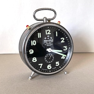 Puede incluir: Un reloj despertador plateado con una esfera negra y números blancos. El reloj tiene un asa en la parte superior y tres patas. La esfera del reloj dice "Wehrle Three-In-One Jeweled Rust & Dust Protected Shockproof".