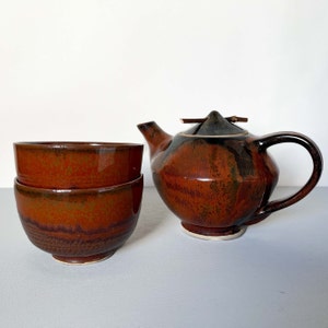 以下が含まれることがあります： 蓋と2つの茶碗のセットが付いた、茶色と黒のセラミック製の急須。急須は独特の、わずかに非対称な形をしています。