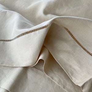 May include: A close-up of a white linen tablecloth with a delicate, stitched border. The fabric is soft and textured, creating a luxurious feel.