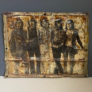 Può includere: Un'insegna in metallo vintage con un'immagine in bianco e nero di una band. L'insegna mostra segni di usura, con ruggine, macchie e pieghe. I membri della band sono disposti in fila, con uno sfondo sbiadito.