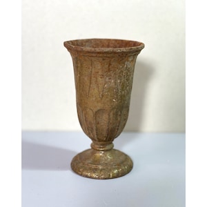 May include: An aged, antique-style vase with a pedestal base. The vase has a fluted body and a wide, slightly flared rim. The colour is a muted gold with areas of rust and green, giving it a vintage appearance. The vase is likely made of metal.