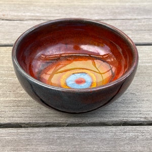 Puede incluir: Un pequeño cuenco de cerámica hecho a mano con un interior marrón rojizo brillante y un exterior oscuro color carbón. El interior del cuenco presenta un diseño abstracto colorido con tonos azules, amarillos y naranjas. El cuenco se utiliza probablemente para servir o exhibir.