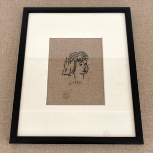 Puede incluir: Obra de arte enmarcada con un marco negro, que presenta un retrato bordado en blanco y negro del perfil de una persona. La obra de arte está sobre un fondo de tela beige, con el nombre "G. FERM" bordado debajo.