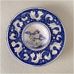 May include: A blue and white ceramic plate with a decorative design of stylized leaves and a central image of a figure in a hat holding a staff. The plate has a raised rim and a slightly indented center.