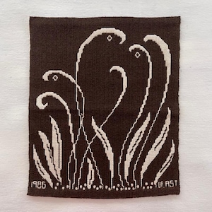 Peut inclure: Textile marron avec un motif floral blanc brodé. Le motif présente des tiges et des feuilles stylisées, avec l'année "1986" et le nom "UF.AST" brodés en bas. Le textile est de forme carrée.