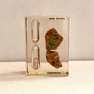 May include: A clear acrylic block containing two pieces of chalcosite, a copper sulfide mineral. The block also features a small hourglass. The label on the block reads "Pierre Veritable Chalcosite".