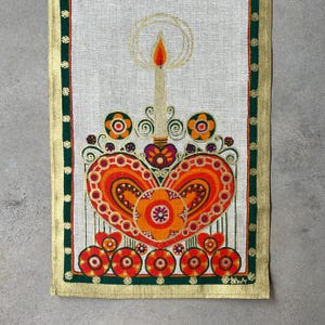 Könnte beinhalten: Ein vintage Tischläufer aus Leinen mit einem farbenfrohen Herzdesign und einer Kerze in der Mitte. Das Herz ist von Blumen und Schnörkeln in Orange-, Rot-, Grün- und Goldtönen umgeben. Der Läufer hat einen goldenen Rand mit einem grün-goldenen Muster.