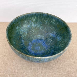 Pode incluir: Uma tigela de cerâmica com formato profundo e arredondado. O interior apresenta uma mistura de tons azuis e verdes, com uma textura salpicada. A borda tem um tom mais escuro e terroso, e a tigela repousa sobre uma superfície neutra.