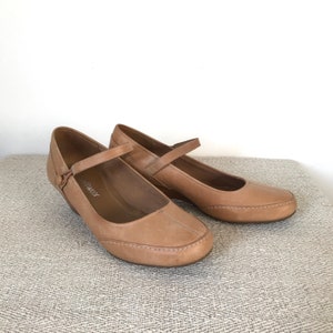 K&ouml;nnte beinhalten: Ein Paar flache Mary Jane-Schuhe aus hellbraunem Leder. Die Schuhe haben eine runde Spitze, einen Riemen &uuml;ber dem Oberteil mit Knopfverschluss und einen kleinen Keilabsatz. Die N&auml;hte sind um die Kanten des Schuhs sichtbar.