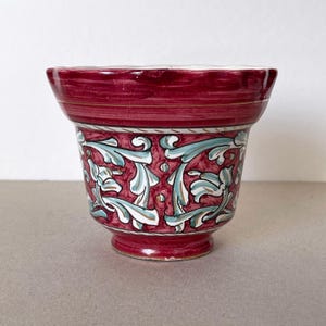 Op de afbeelding: Een bordeauxrode keramische bloempot met een uitlopende rand en een afgeronde basis. De bloempot heeft turquoise en witte bloempatronen. Gouden details zijn aanwezig bij de rand. Geschikt voor binnen of buiten. Afmetingen in centimeters.