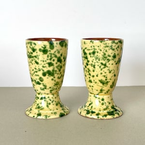 Puede incluir: Dos tazas de cerámica amarilla con esmalte verde moteado. Las tazas tienen una base ancha y una parte superior estrecha.