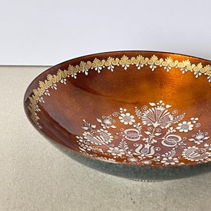 Könnte beinhalten: Eine braune Keramikschale mit weißen und goldenen bemalten Blumendesigns. Die Schale hat eine glänzende Oberfläche und einen leicht ausgestellten Rand.