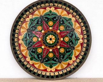 May include: A round brass plate with a colorful floral design. The plate features a central starburst pattern with red, yellow, green, and black accents. The outer rim of the plate is decorated with a black and gold border.