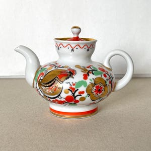 Puede incluir: Tetera de cerámica blanca con tapa, decorada con un diseño floral y de pájaros en dorado, rojo y verde. La tetera tiene un pico curvo y un asa redondeada. La tapa es roja con un pomo dorado.