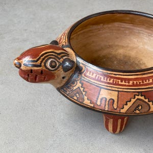 May include: A ceramic bowl shaped like a turtle with a wide, open top. The exterior features hand-painted geometric patterns in shades of terracotta, cream, and black. The bowl is supported by three short, sturdy legs and has a sculpted head.