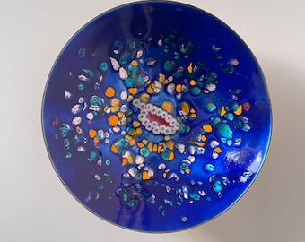 Könnte beinhalten: Eine blaue Keramikschale mit einem farbenfrohen, abstrakten Design. Die Schale hat einen zentralen Cluster aus gelben, weißen und grünen Formen, umgeben von kleineren, verstreuten Formen.