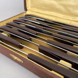 May include: A set of 12 vintage knives with dark brown handles and silver blades, arranged in a brown leather case.