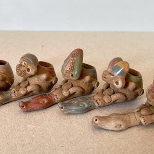 Puede incluir: Una colección de pequeñas figuras de arcilla hechas a mano. Cada figura presenta una forma humanoide sentada con una abertura en forma de anillo. Las figuras están pintadas con acentos rojos, marrones y verdes. Las figuras están dispuestas en fila.