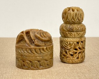 May include: Two small, intricately carved, brown stone boxes. One box is round with a floral design and the other is cylindrical with a stacked design.