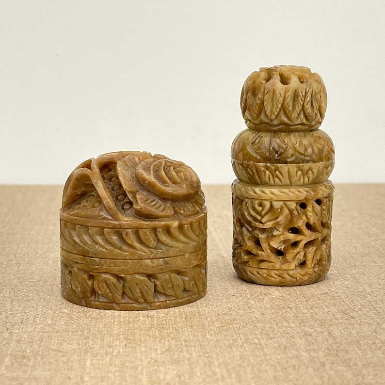Puede incluir: Dos peque&ntilde;as cajas de piedra marr&oacute;n, talladas de forma intrincada. Una caja es redonda con un dise&ntilde;o floral y la otra es cil&iacute;ndrica con un dise&ntilde;o apilado.
