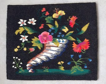 Op de afbeelding: Een zwart geweven wandkleed met een kleurrijk bloemenmotief. De bloemen zijn gerangschikt in een hoorn des overvloeds, wat een symbool is van overvloed en welvaart. Het wandkleed is een prachtig voorbeeld van traditionele volkskunst.