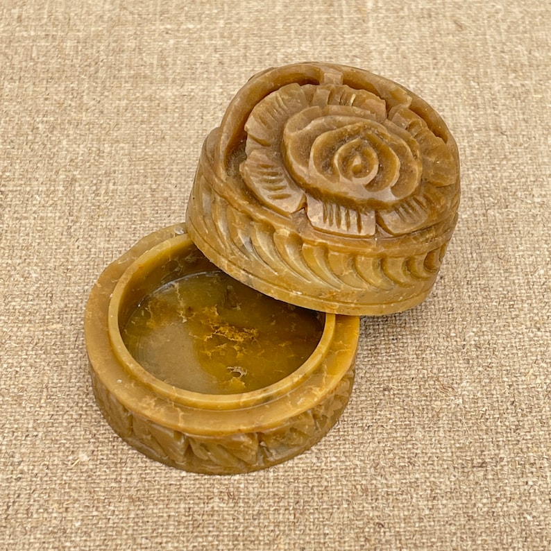 Puede incluir: Una peque&ntilde;a caja de piedra de color marr&oacute;n amarillento tallada con un dise&ntilde;o floral. La tapa est&aacute; abierta y el interior de la caja es visible.