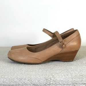 K&ouml;nnte beinhalten: Ein Paar hellbraune Leder-Keilabsatzschuhe mit Riemen und Knopfverschluss. Die Schuhe haben eine runde Spitze und einen Holzkeilabsatz. Die Schuhe sind etwa 7.6 cm hoch.