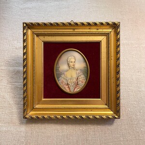 Pode incluir: Um retrato em miniatura emoldurado de uma mulher em uma moldura oval. O retrato apresenta uma mulher em um vestido rosa e branco, com uma touca de renda branca e um colar de pérolas. A moldura é dourada com um design de corda.