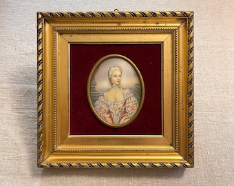 May include: A framed miniature portrait of a woman in an oval frame. The portrait features a woman in a pink and white dress, with a white lace cap and a pearl necklace. The frame is gold with a rope design.